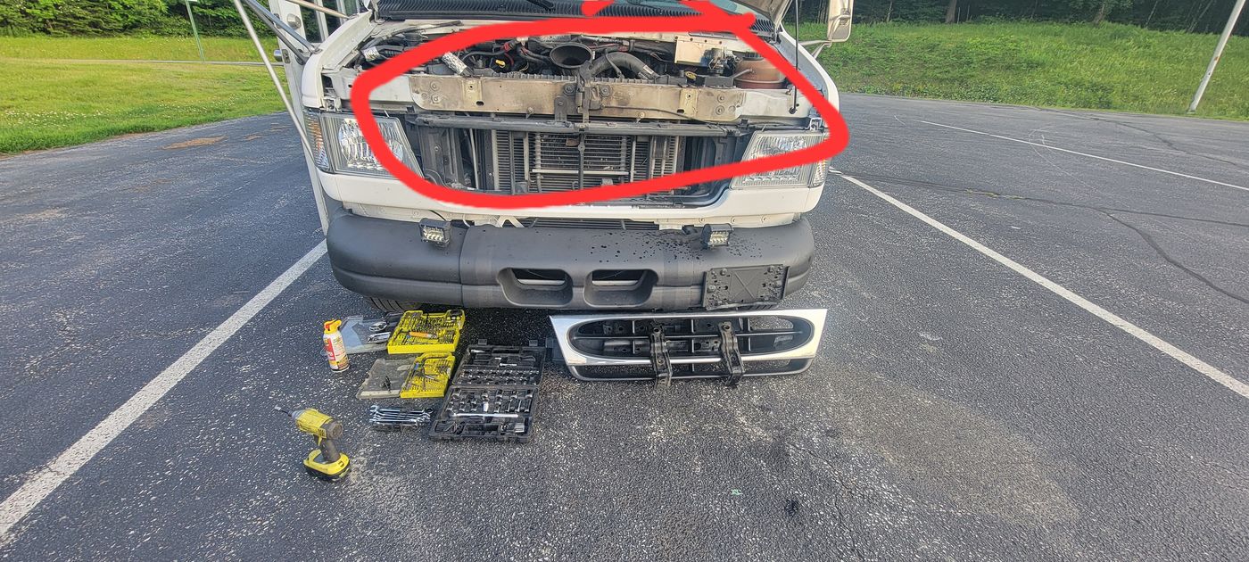 Front grille removed for engine bay access, radiator and hoses exposed, tools laid out on the ground below.