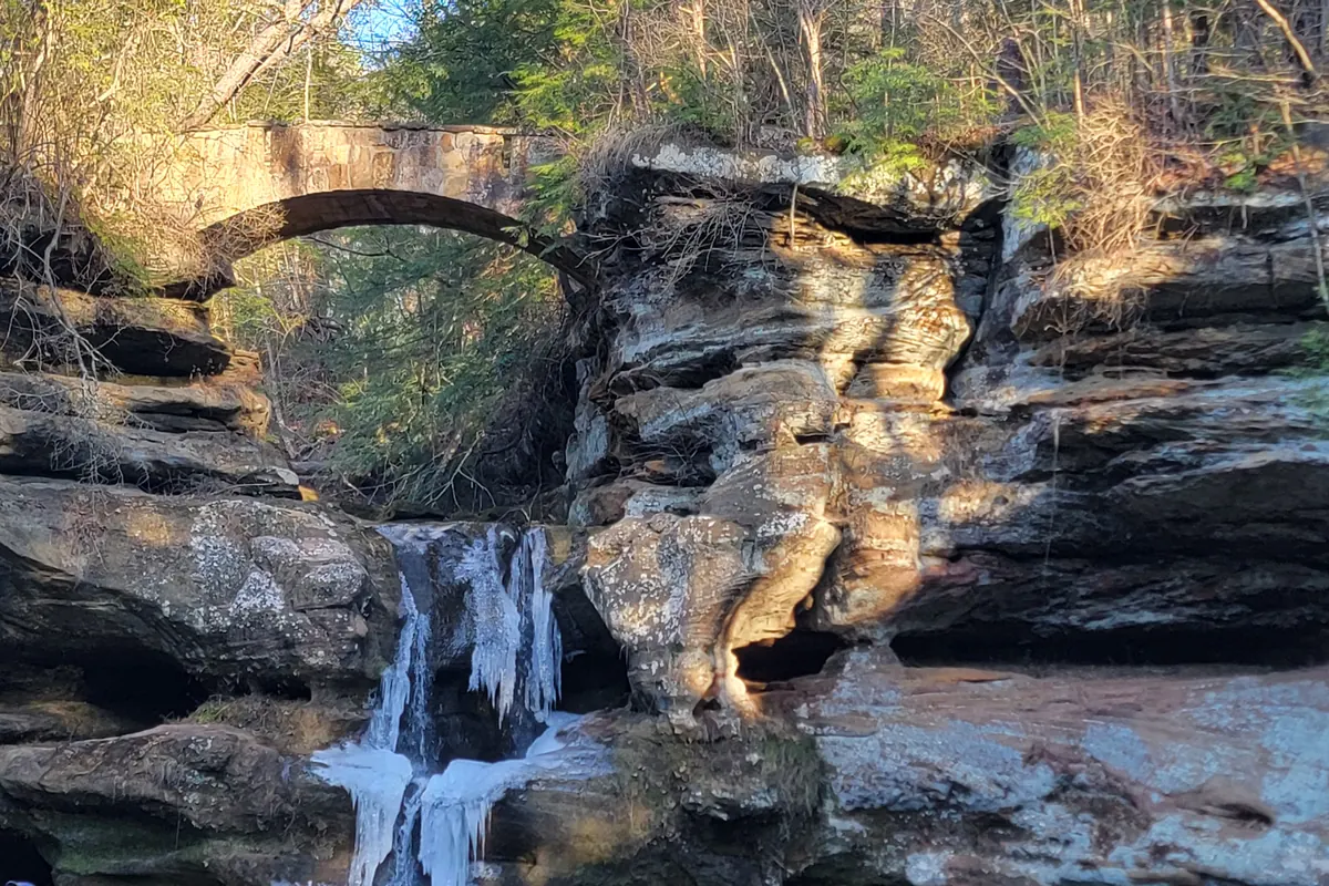 A partly frozen waterfall below a stone arch bridge in a sandstone gorge